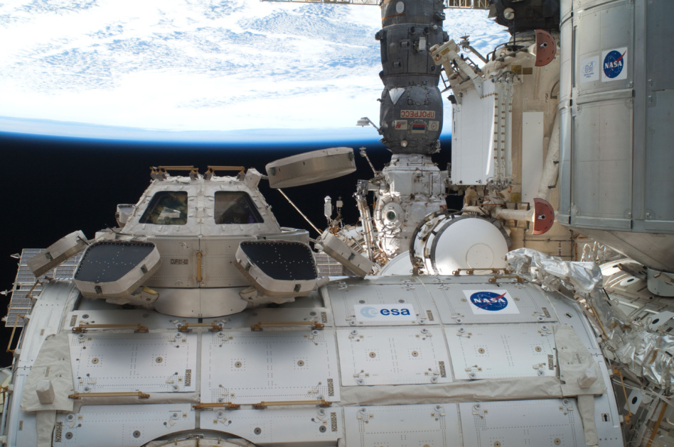 The International Space Station's Cupola, backdropped against black space, a horizon scene array and various components of the orbiting outpost, including Node 3 or Tranquility (on which the Cupola is mounted) and the Leonardo Permanent Multipurpose Module.