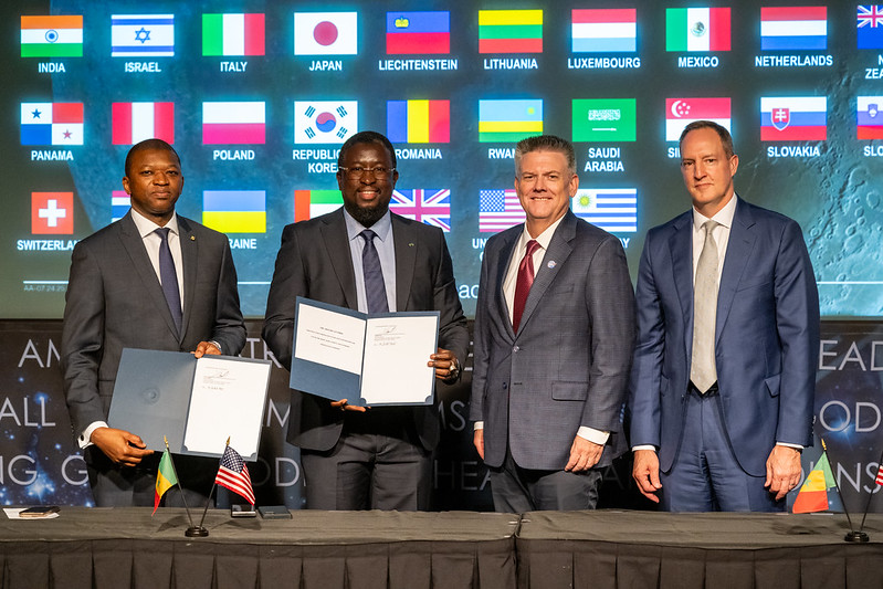 Four men in suits hold up signed documents in front of a wall of flags